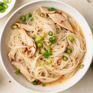 A close-up of chicken long rice recipe featuring clear bean thread noodles, tender shredded chicken, and sliced green onions in a savory broth, served in a speckled ceramic bowl.