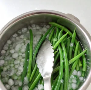 vibrant green beans submerged in a stainless steel bowl filled with an ice water bath, with metal tongs resting on top to help blanch them.