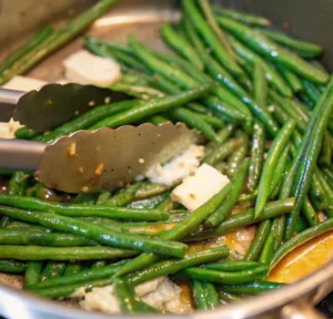 Close-up of french green beans being sautéed in a pan with melting pats of butter and minced garlic, as metal tongs toss them to create a glossy, flavorful glaze.