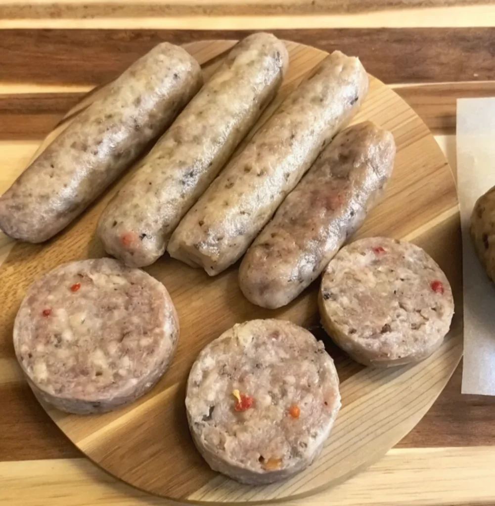 Four uncooked, pale Irish sausages and three thick sausage patties are arranged on a circular wooden board, displaying a smooth texture with visible specks of herbs and red spices.