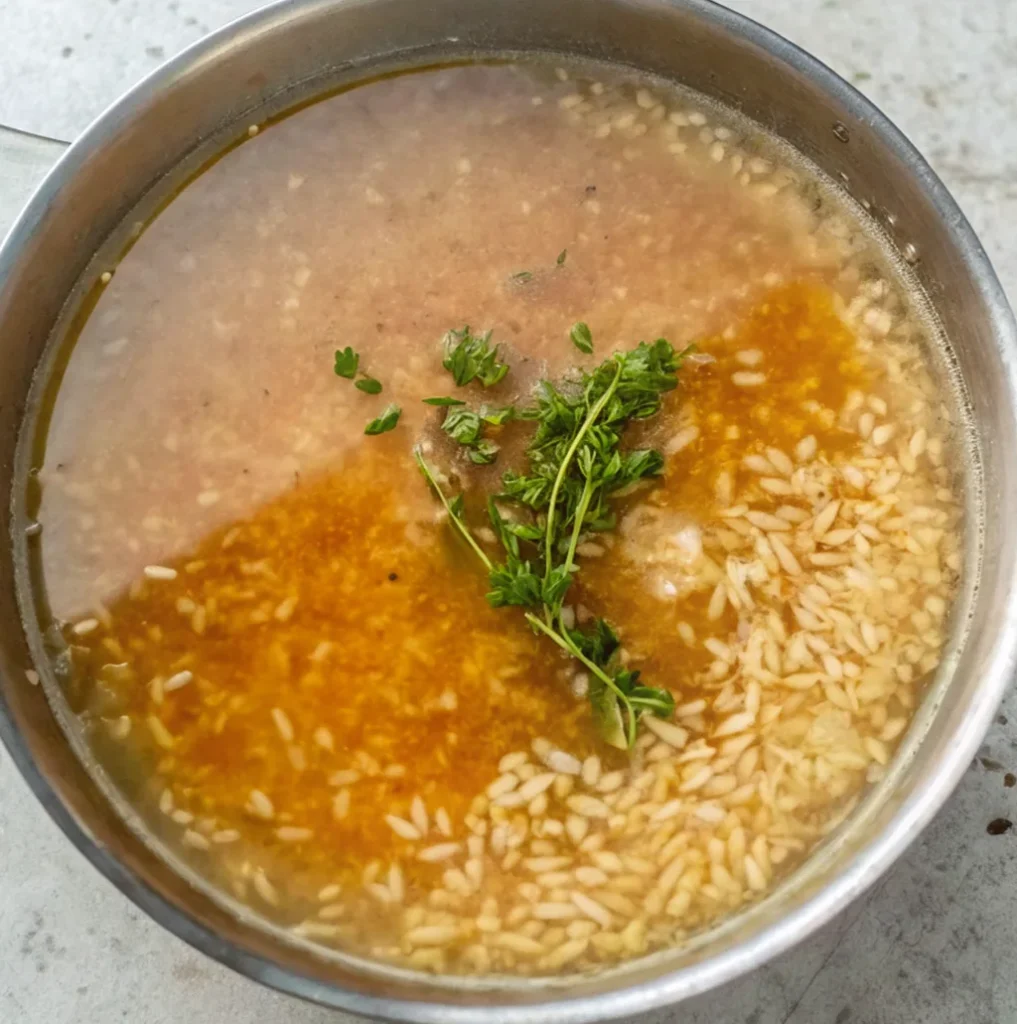 The rice is submerged in chicken broth in the pan. A sprig of fresh green herbs, such as thyme, is placed on top of the rice to add flavor.