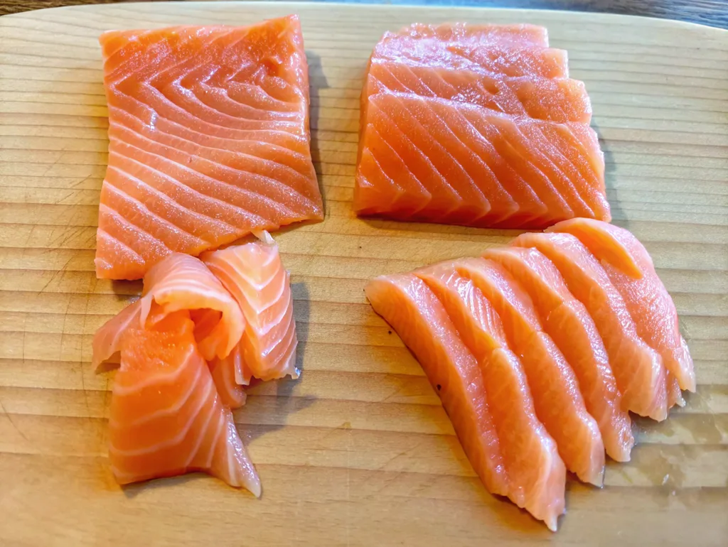 Fresh, vibrant orange salmon sashimi slices with distinct white marbling are displayed on a light wooden cutting board. The arrangement includes thick rectangular cuts and delicate, fanned slices ready for serving.