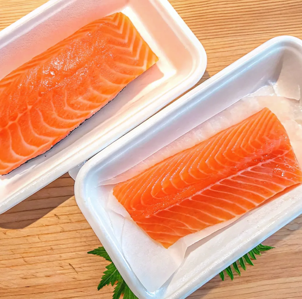 Two fresh, vibrant orange salmon fillets with distinct white marbling are presented in white rectangular trays.