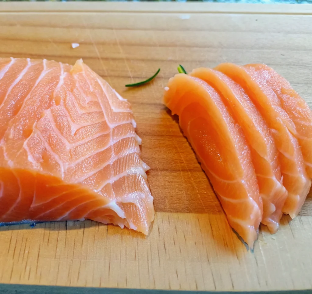 A close-up of a fresh salmon fillet and several thick, orange sashimi slices showing white marbling, resting on a wooden cutting board with a small green garnish.