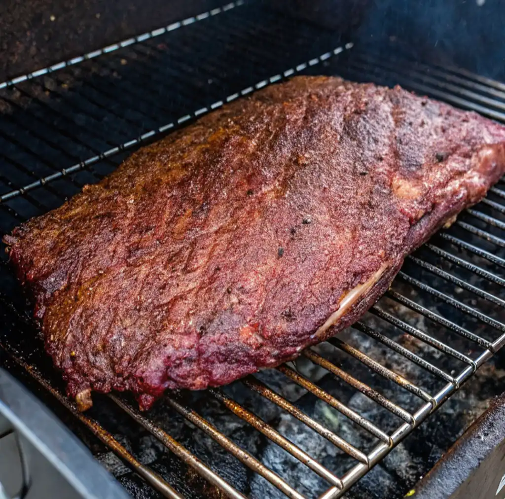 A close-up shot of a seasoned, beef brisket resting on a metal grill grate over charcoal, beginning to smoke and develop a rich, mahogany-colored exterior bark.