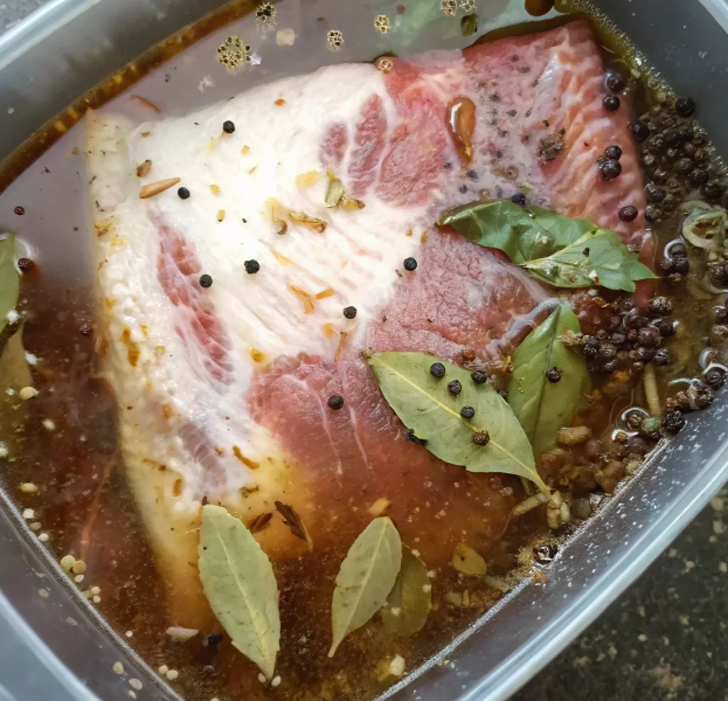 Brining Method: A raw brisket submerged in a dark liquid brine within a clear container, surrounded by aromatic whole peppercorns and bay leaves to enhance the meat's tenderness before cooking.