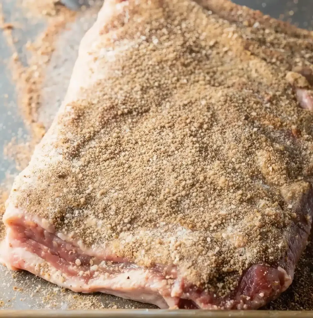 A raw brisket is shown being heavily coated with a coarse dry brine rub consisting of salt, pepper, and spices, which covers the entire surface to build flavor.