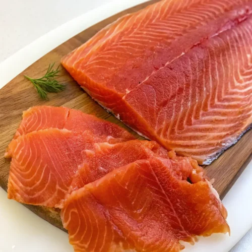 A vibrant, orange-red fillet of cold-smoked steelhead trout rests on a wooden board. Several thin, delicate slices are layered in the foreground, showcasing the fish's glossy texture and white marbling.