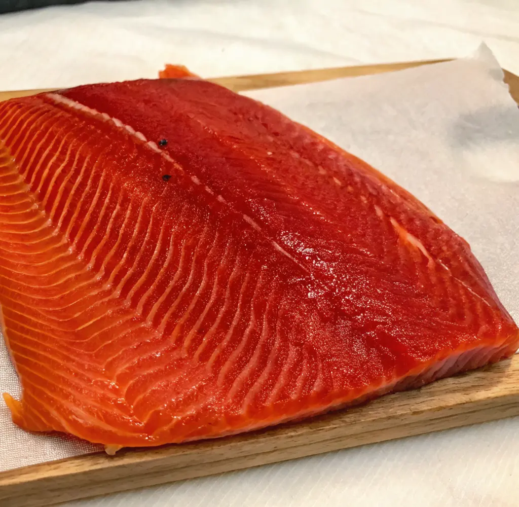A thick, glossy fillet of cold-smoked steelhead trout displays a deep ruby-red color and distinct white fat lines. It rests on parchment paper atop a rustic wooden serving board.