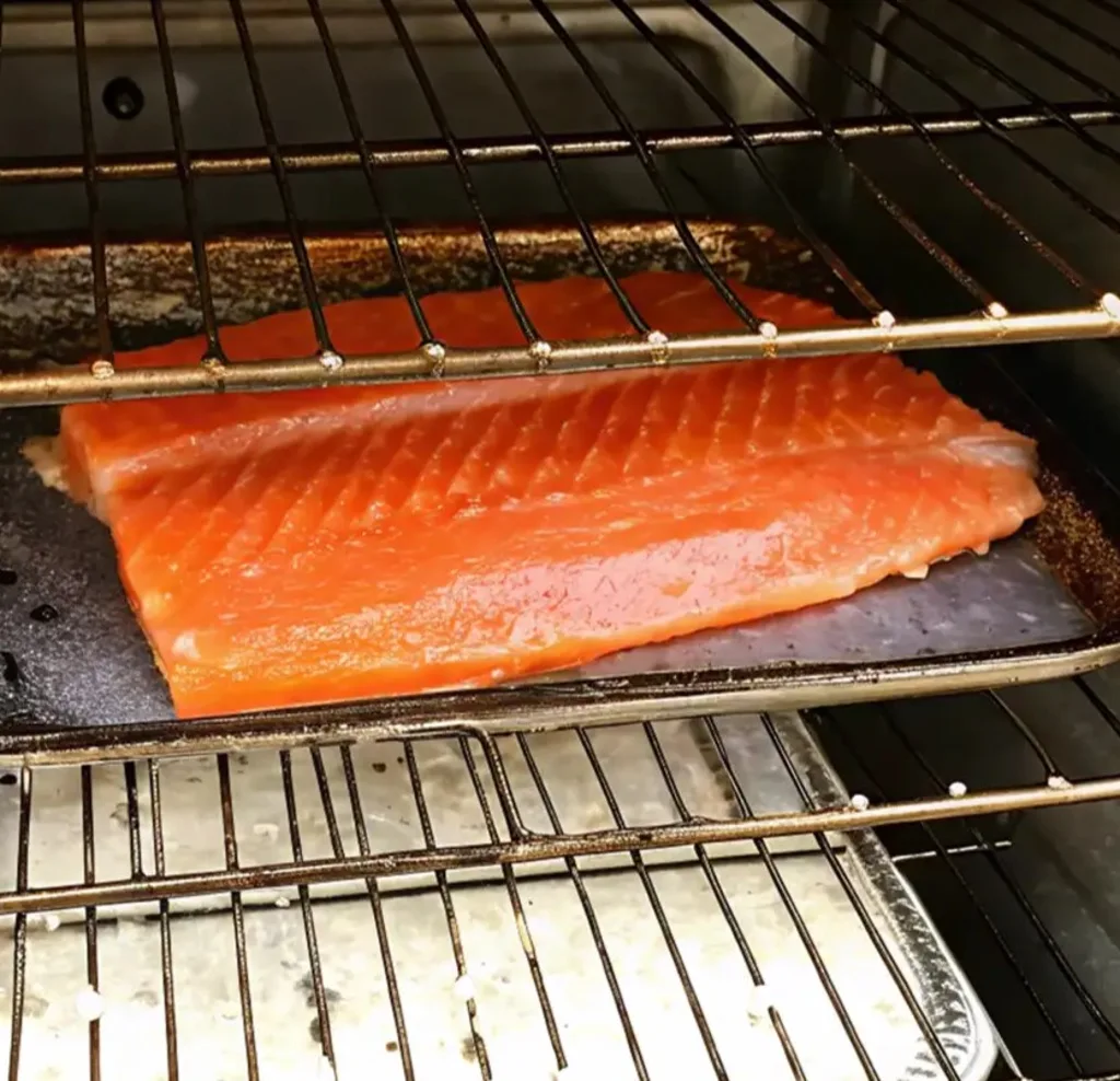 A vibrant, deep-orange steelhead trout fillet sits on a wire rack inside a smoker. A second tray rests below it, capturing drippings during the cold-smoking process for a rich flavor.