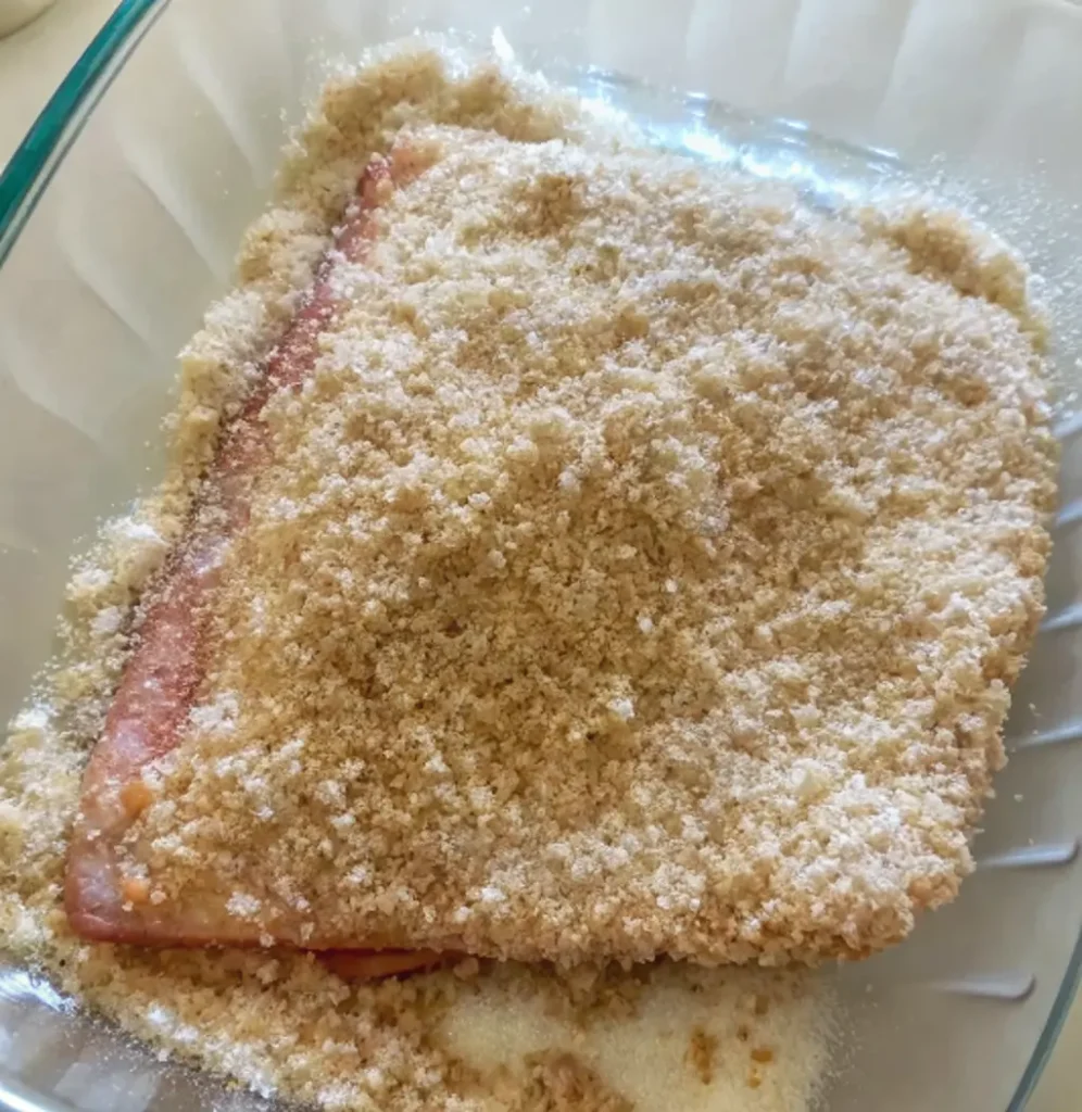 A vibrant, pink steelhead trout fillet is completely blanketed in a coarse, sandy-textured dry cure of salt and sugar within a glass dish to draw out moisture before smoking.