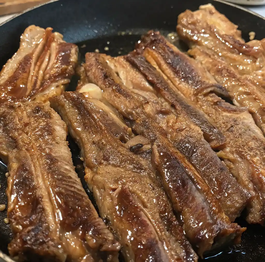 The beef short ribs are flipped in the skillet, showing a deep golden-brown crust and rendered fat as they continue to cook evenly across the pan’s surface.