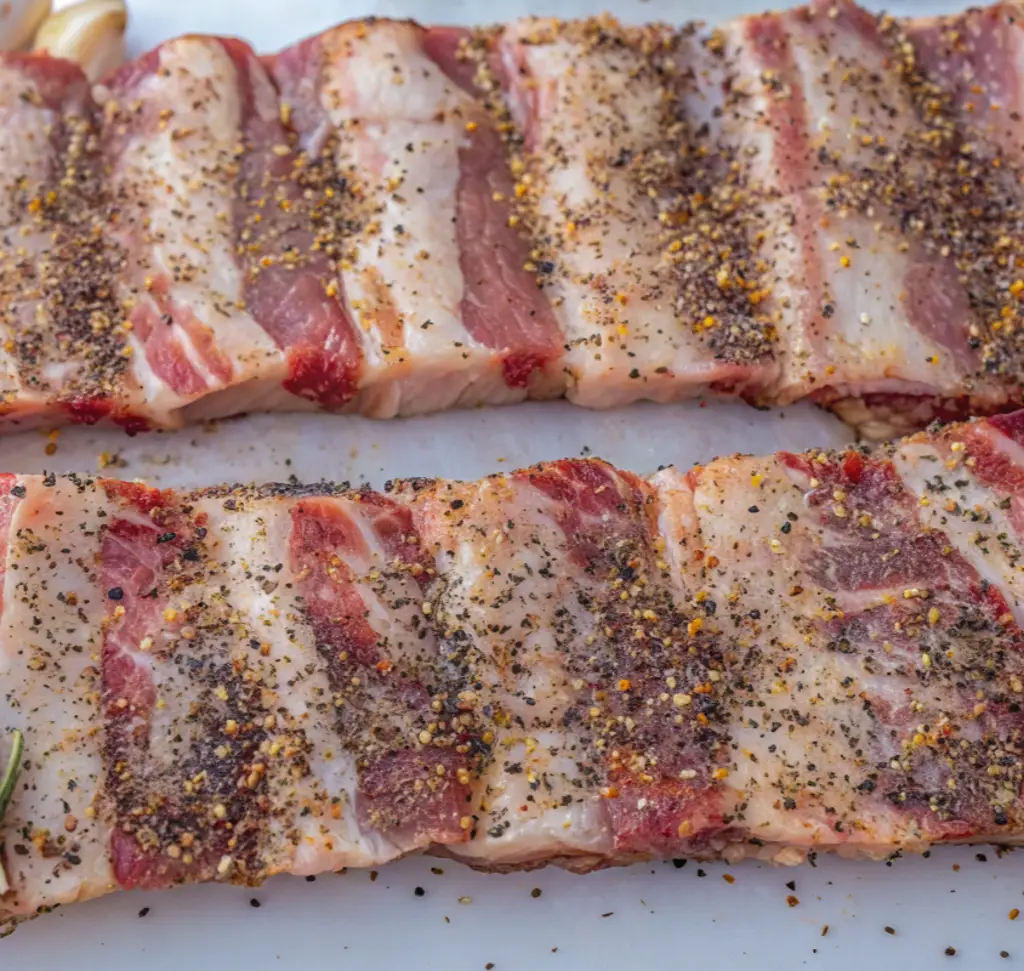 Two long slabs of raw, marbled beef short ribs are laid flat and generously coated with a coarse dry rub of salt, black pepper, and garlic on a white surface.
