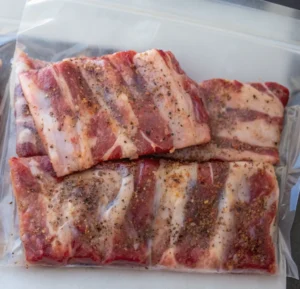 Seasoned raw beef short ribs are neatly packed inside a clear vacuum-seal plastic bag, ready for the sous-vide water bath, with the dry rub visible on the meat.
