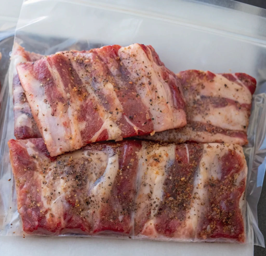 Seasoned raw beef short ribs are neatly packed inside a clear vacuum-seal plastic bag, ready for the sous-vide water bath, with the dry rub visible on the meat.