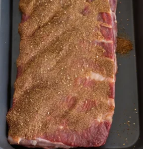 Raw beef ribs laid in a dark roasting pan, generously coated with a savory brown spice rub. The meat shows marbled texture, prepared and ready for baking at 350°F.