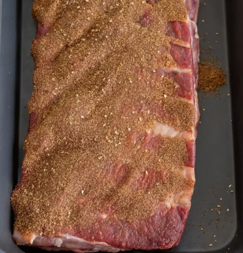 Raw beef ribs laid in a dark roasting pan, generously coated with a savory brown spice rub. The meat shows marbled texture, prepared and ready for baking at 350°F.