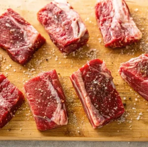 Six thick cuts of raw boneless beef ribs seasoned with coarse sea salt and cracked black pepper, arranged on a wooden cutting board during the meal preparation process.