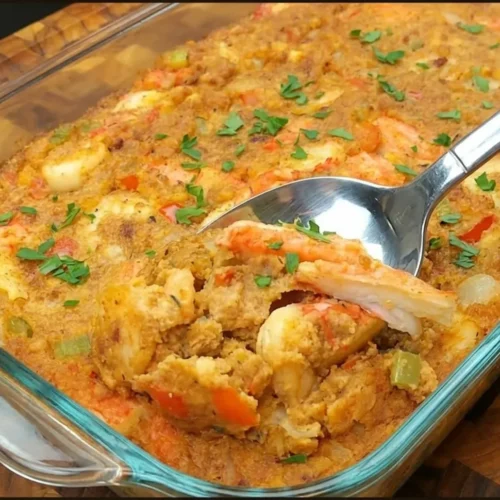 A large silver spoon scoops a serving of moist cornbread seafood dressing from a glass dish, highlighting tender shrimp, crab meat, and a fresh garnish of chopped parsley.
