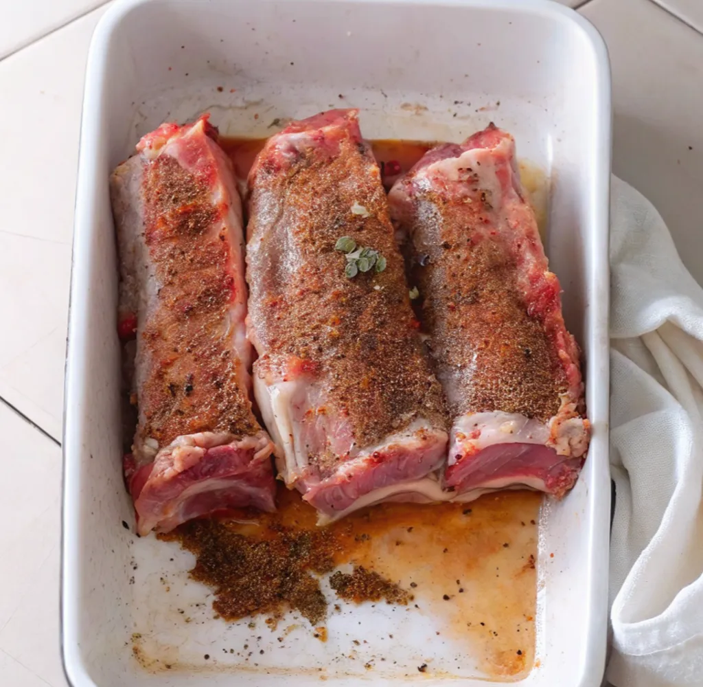 Three beef short ribs arranged in a white ceramic baking dish, coated in a thick layer of dry rub seasoning and aromatics, prepared for slow roasting or braising.