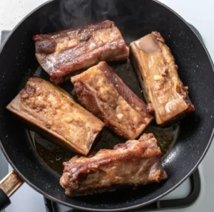 Beef short ribs being seared in a hot black skillet until golden brown, with steam rising and natural juices sizzling, to lock in flavor before the braising process.