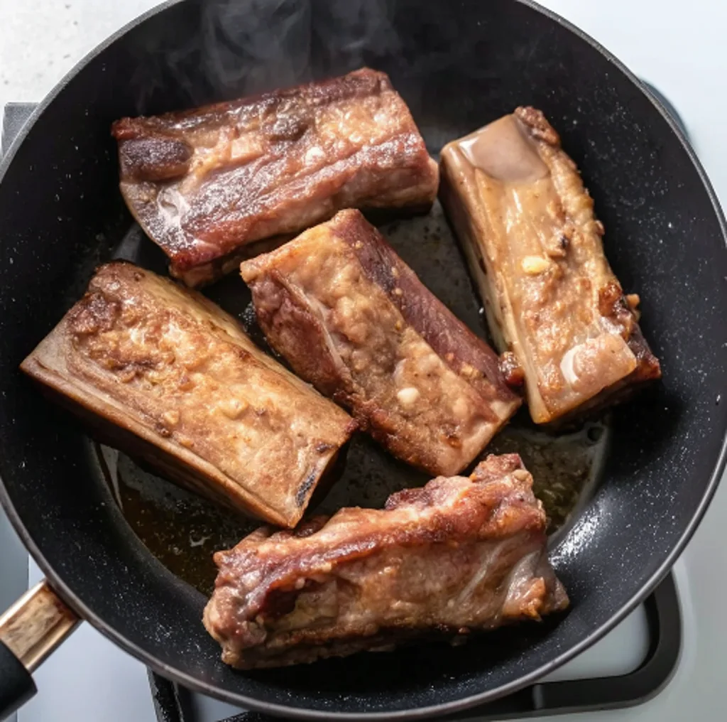 Beef short ribs being seared in a hot black skillet until golden brown, with steam rising and natural juices sizzling, to lock in flavor before the braising process.