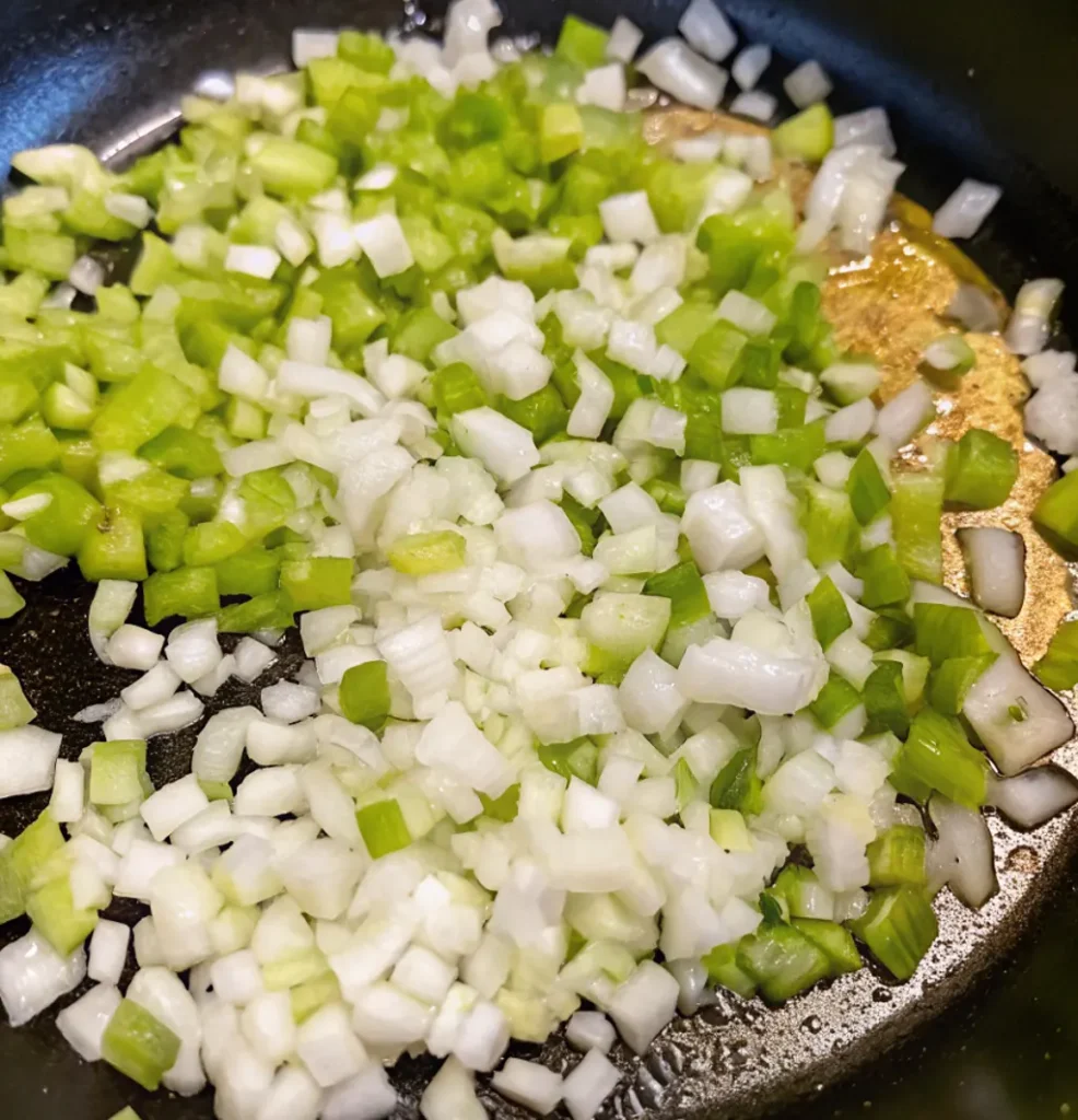 Finely diced white onions and green celery sautéing in a hot pan with oil. The vegetables are crisp and bright, forming the aromatic base for the seafood dressing recipe