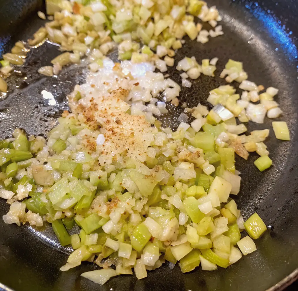 A close-up shot of chopped onions and celery softening in a black skillet. This essential cooking step builds the flavor foundation for a traditional oyster dressing dish.