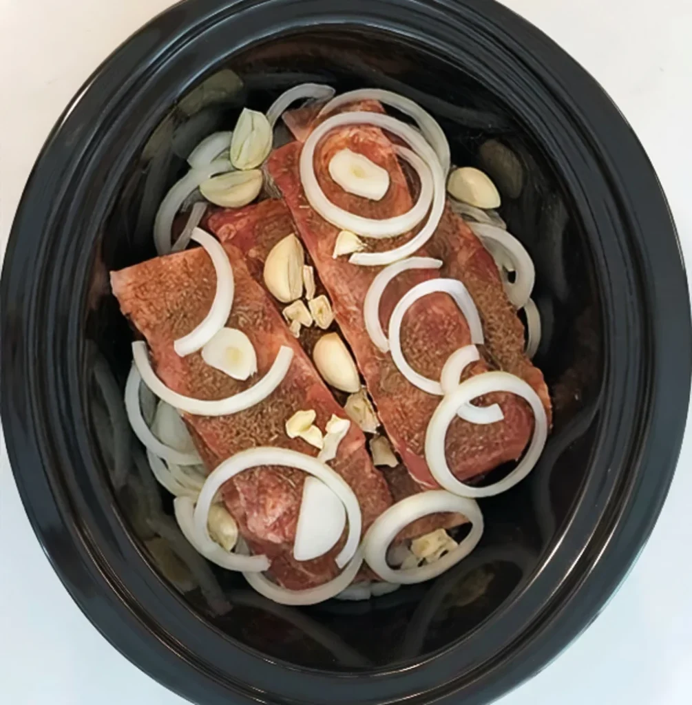 Raw beef ribs seasoned with spices and layered with fresh white onion rings and whole garlic cloves inside a black slow cooker