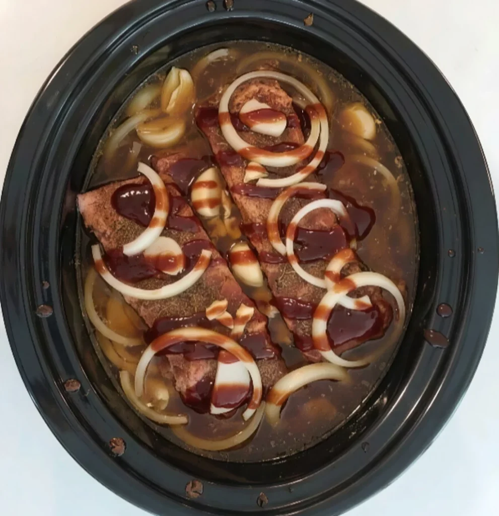 Seasoned beef ribs in a slow cooker submerged in a savory broth, topped with sliced onions, garlic cloves, and a rich barbecue sauce drizzle