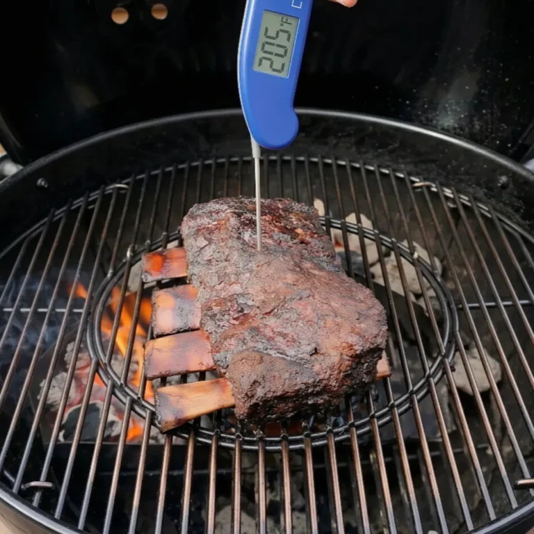 A digital meat thermometer displays an internal temperature of 205°F as it probes a thick, smoked rack beef ribs internal temperature of beef ribs resting on a charcoal grill