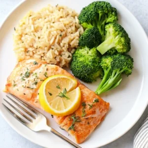 A beautifully plated meal featuring a flaky Baked Salmon Recipe fillet topped with a lemon slice and herbs, served alongside a portion of seasoned rice and vibrant steamed broccoli florets.