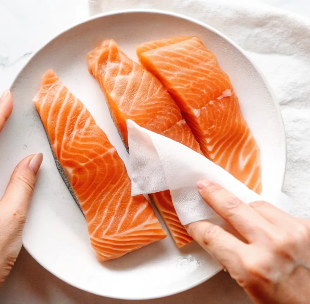 white paper towel to pat dry three fresh, vibrant orange raw salmon fillets arranged on a light grey plate