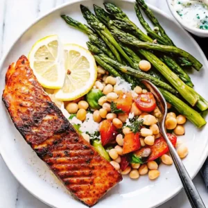 A beautifully seared Grilled Salmon fillet is served with charred asparagus, a vibrant chickpea and tomato salad, and fresh lemon slices
