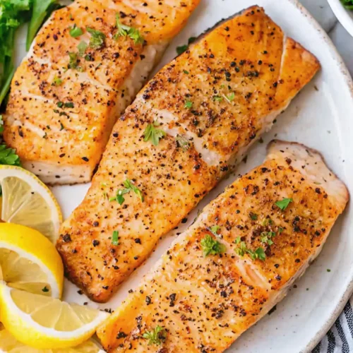 Golden, Pan Seared Salmon fillets seasoned with cracked black pepper and fresh herbs, served on a white plate with bright lemon slices and parsley garnish