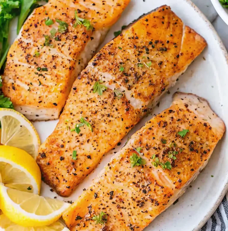 Golden, Pan Seared Salmon fillets seasoned with cracked black pepper and fresh herbs, served on a white plate with bright lemon slices and parsley garnish