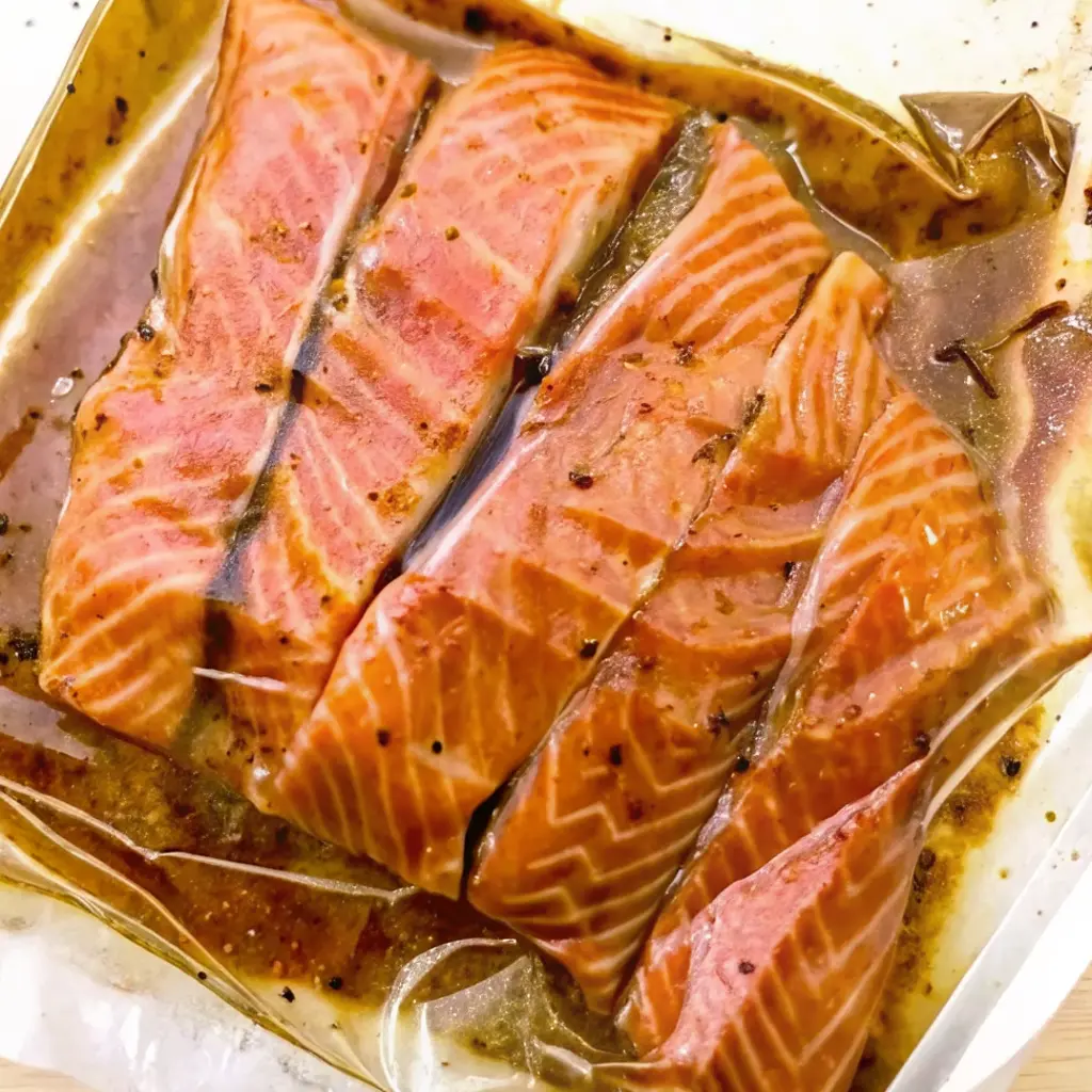 Fresh salmon fillets soaking in a rich, savory Salmon Marinade within a bag. The image highlights the seasoning specks and the glossy texture of the fish as it infuses.