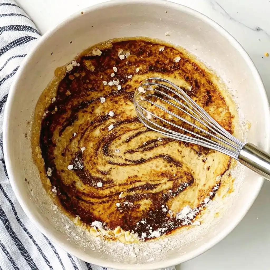 A stainless steel whisk mixing a dark, savory Salmon Marinade with light spices in a white ceramic bowl.