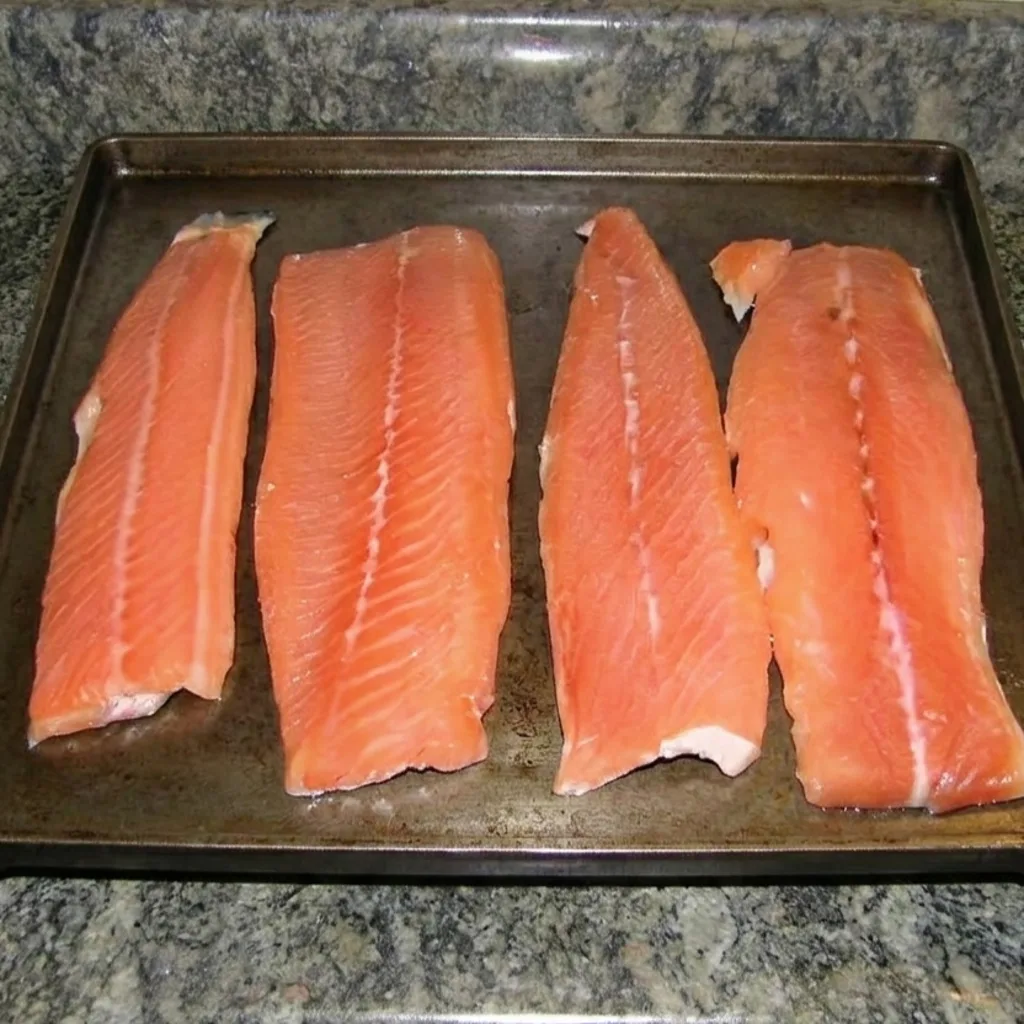 Four raw salmon fillets rest on a dark baking sheet atop a granite countertop, ready for brining and smoking.
