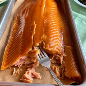 A large, beautifully for Smoked Salmon Brine Recipe in a metal tray, its golden-orange surface glistening. A fork pulls away tender, succulent pieces, revealing the moist, perfectly cured interior texture.