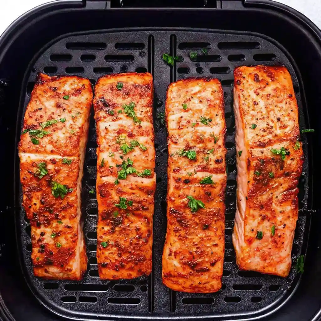 Four perfectly golden-brown air fryer salmon topped with fresh parsley, resting on a black air fryer tray.