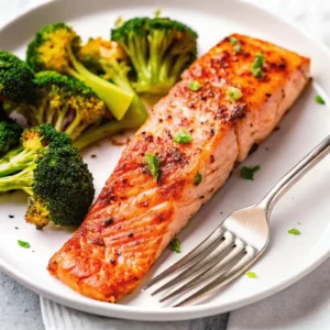 A single air fryer salmon fillet with a crispy, seasoned crust served alongside roasted broccoli florets on a white plate, offering a healthy and balanced low-carb meal idea for dinner.