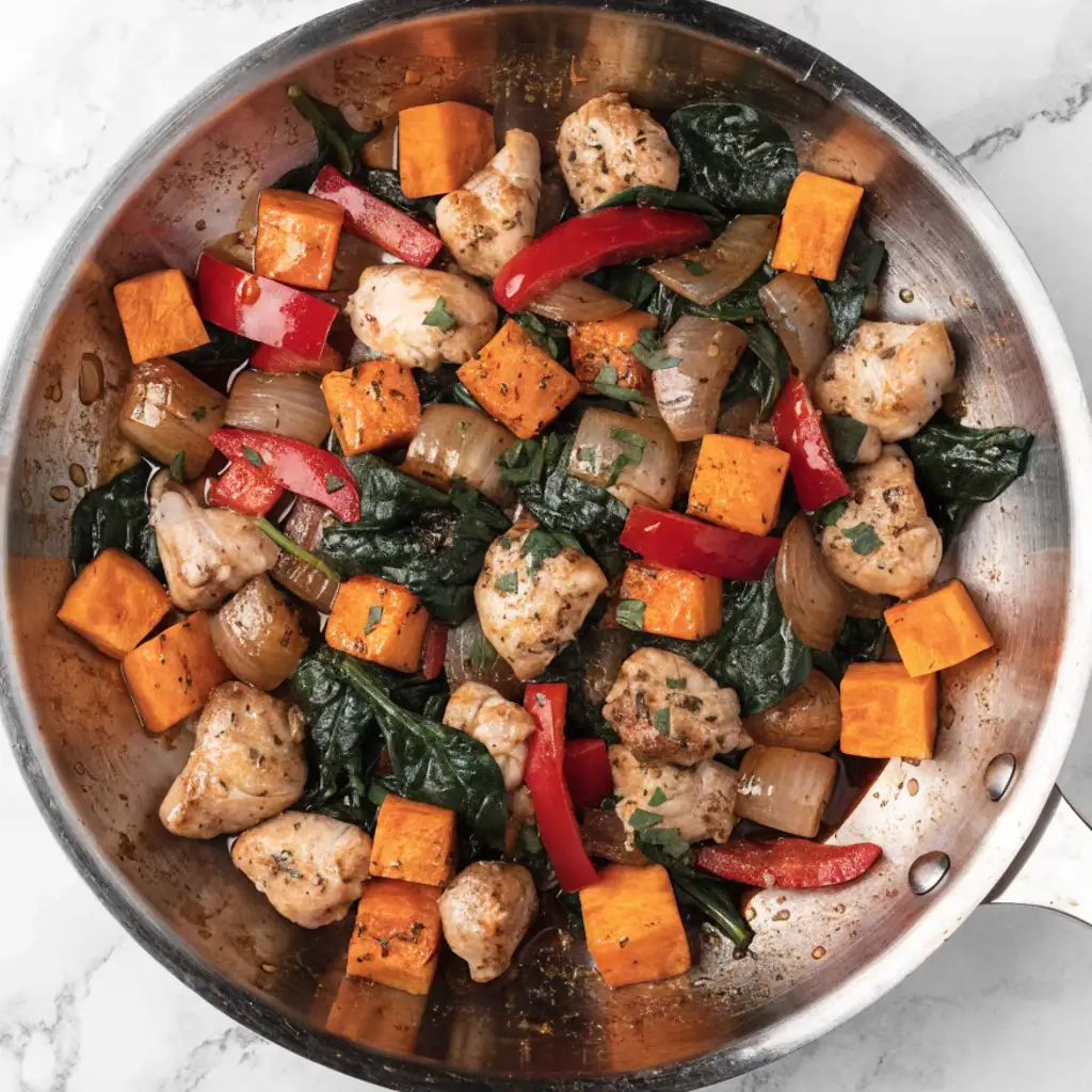 A top-down view of a stainless steel skillet filled with a healthy chicken Recipes featuring tender chicken breast chunks, cubed sweet potatoes, red bell peppers, onions, and wilted spinach.