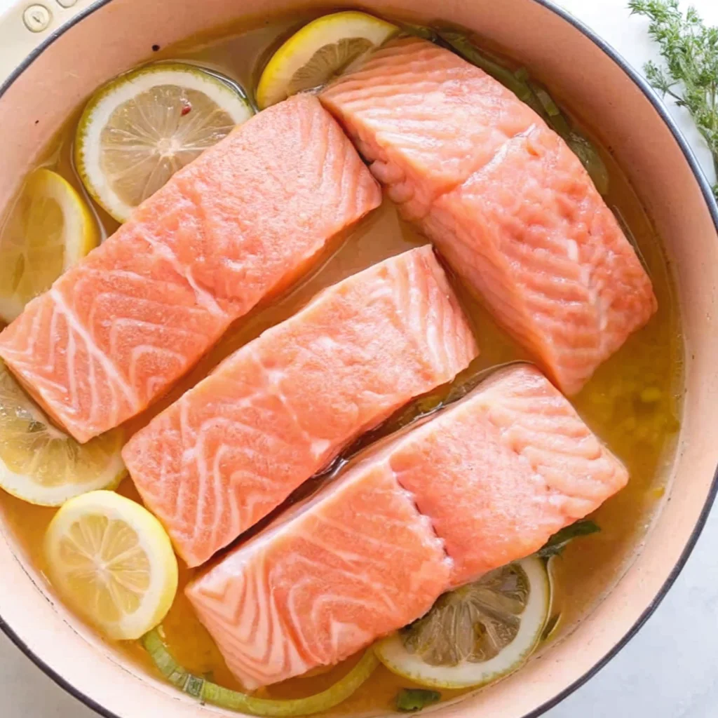 Four salmon fillets poach gently in a shallow pan of golden, citrus-infused broth. Vibrant lemon slices and fresh green herbs surround the fish