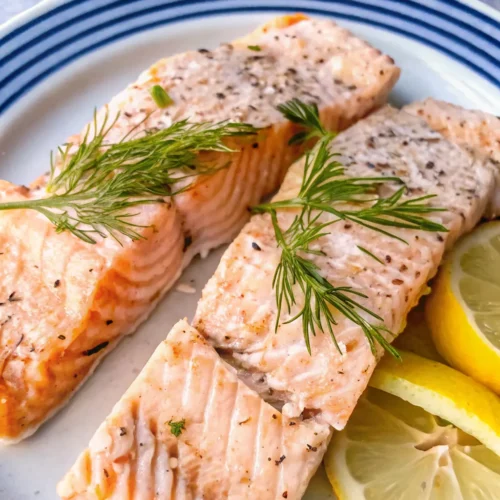 Two tender, poached salmon Recipe fillets are topped with fresh dill sprigs and served with bright lemon slices. The seasoned fish is beautifully flakey and presented on a blue-rimmed white plate.