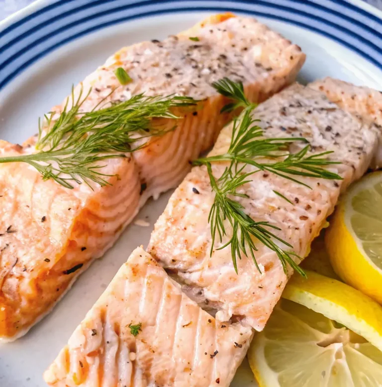 Two tender, poached salmon Recipe fillets are topped with fresh dill sprigs and served with bright lemon slices. The seasoned fish is beautifully flakey and presented on a blue-rimmed white plate.