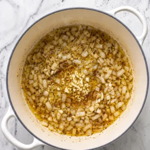 Chopped onions and minced garlic are sautéed in oil in a Dutch oven placed on a marble surface.