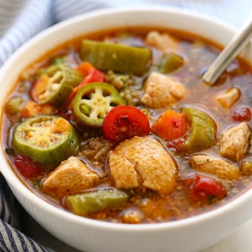 A white bowl filled with hearty chicken gumbo soup, featuring tender chicken chunks, sliced okra, cherry tomatoes, and green bell peppers in a flavorful, seasoned broth with a spoon.