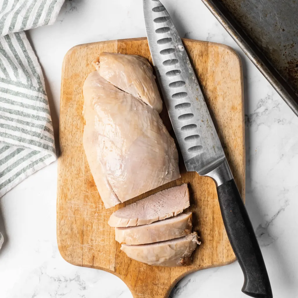 Cooked chicken breast sliced on a wooden cutting board next to a large chef's knife, prepared for Grandma's Chicken Noodle Soup Recipe.
