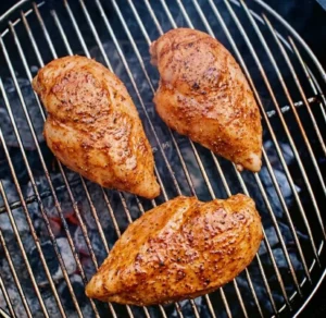 Three seasoned chicken breasts searing on a round charcoal grill grate with visible grill marks, preparing protein for a grilled chicken pasta recipe.